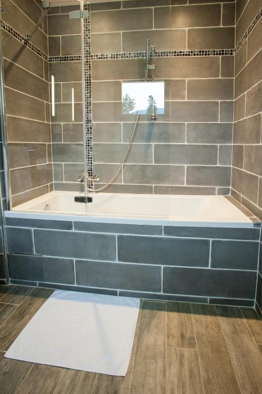 Mūsdienīgs vannas istabas interjers ar pelēkām flīzēm un dušas kabīni, kurā ir vannas un dušas apvienojums. Grīda ir izklāta ar koka izskata flīzēm, radot siltu atmosfēru.