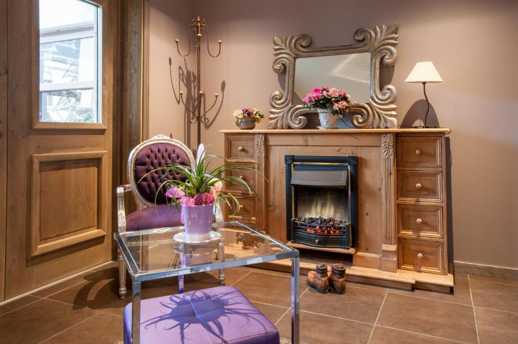 Elegants interjera stūrītis ar purpursarkanu krēslu, stikla galdu un koka kamīnu. Uz kamīna atrodas spogulis un ziedu kompozīcija, radot mājīgu atmosfēru.