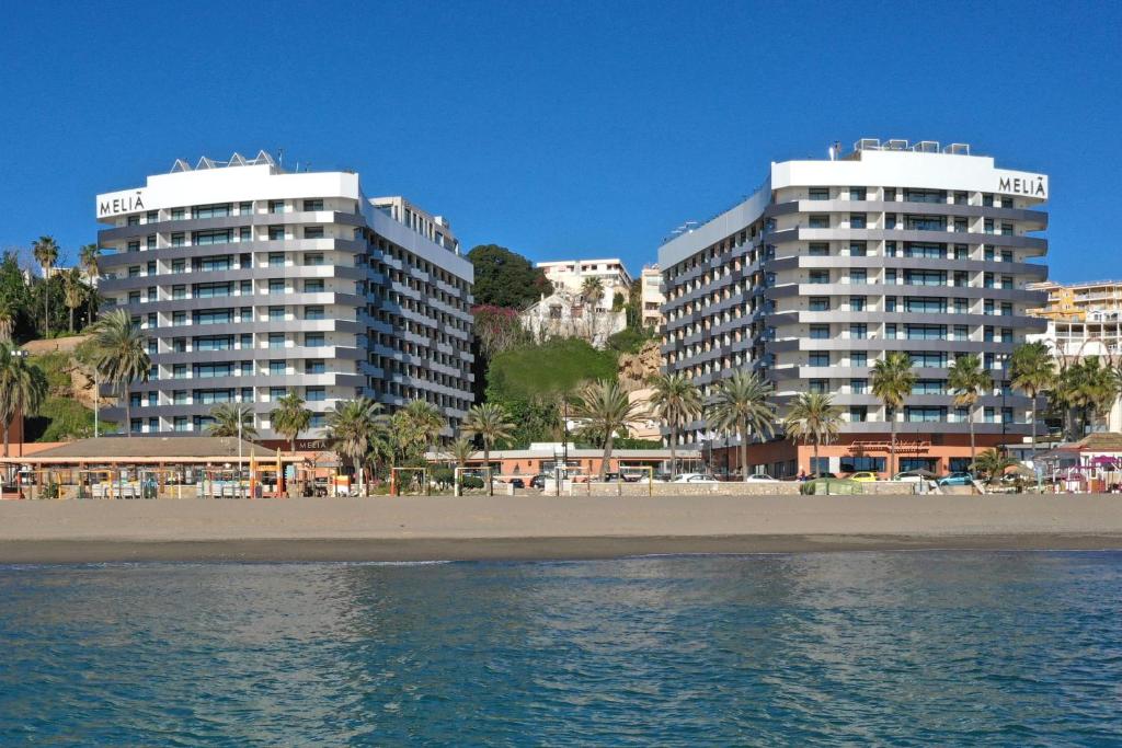 Divi moderni viesnīcu kompleksi pie pludmales ar palmām un skaidru zilu debesi. Viesnīcu nosaukums ir "Meliá".