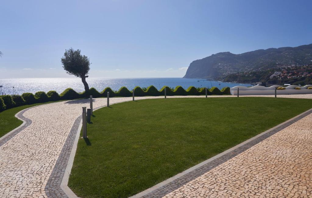 Skats uz jūras krastu ar zaļu zālienu un akmens celiņu, kas ved uz okeānu un kalniem. Saulains laiks un mierīga atmosfēra.