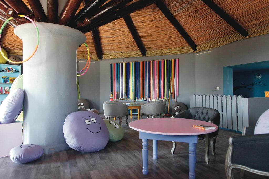 Krāsains bērnu spēļu istabas interjers ar mīkstajiem krēsliem un jautru izskatu. Sienas rotā krāsainas lentes, bet centrā ir apaļa galda ar rozā virsmu.