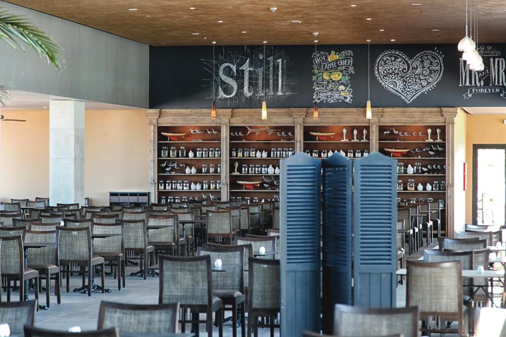 Mūsdienīga restorāna iekšiene ar koka grīdu, izkārtotiem galdiem un krēsliem, un ar mākslinieciskiem uzrakstiem uz sienas. Sienā ir vitrīna ar traukiem un dekoratīviem priekšmetiem.