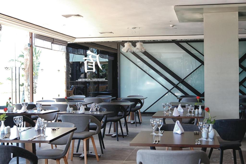 Mūsdienīgs restorāns ar elegantiem galdiem un krēsliem, kur katrs galds ir rotāts ar ziediem un traukiem. Interjerā ir redzams stiklots elements ar ģeometriskām līnijām, kas piešķir modernu noskaņu.