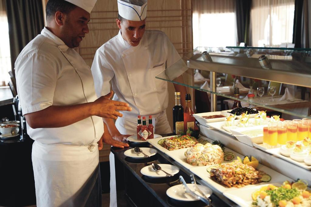 Šefpavāri apspriež ēdienu piedāvājumu, stāvot pie bufetes ar dažādiem ēdieniem un dzērieniem. Galda izkārtojums ir elegants, un redzami daudzi krāsaini ēdieni un saldumi.