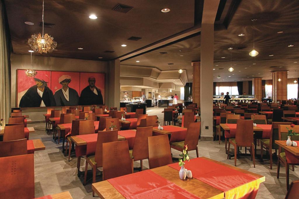 Mūsdienīgs restorāns ar siltu apgaismojumu, koka galdiem un sarkaniem galdautiem, izrotāts ar māksliniecisku gleznu un ziediem uz galdiem.