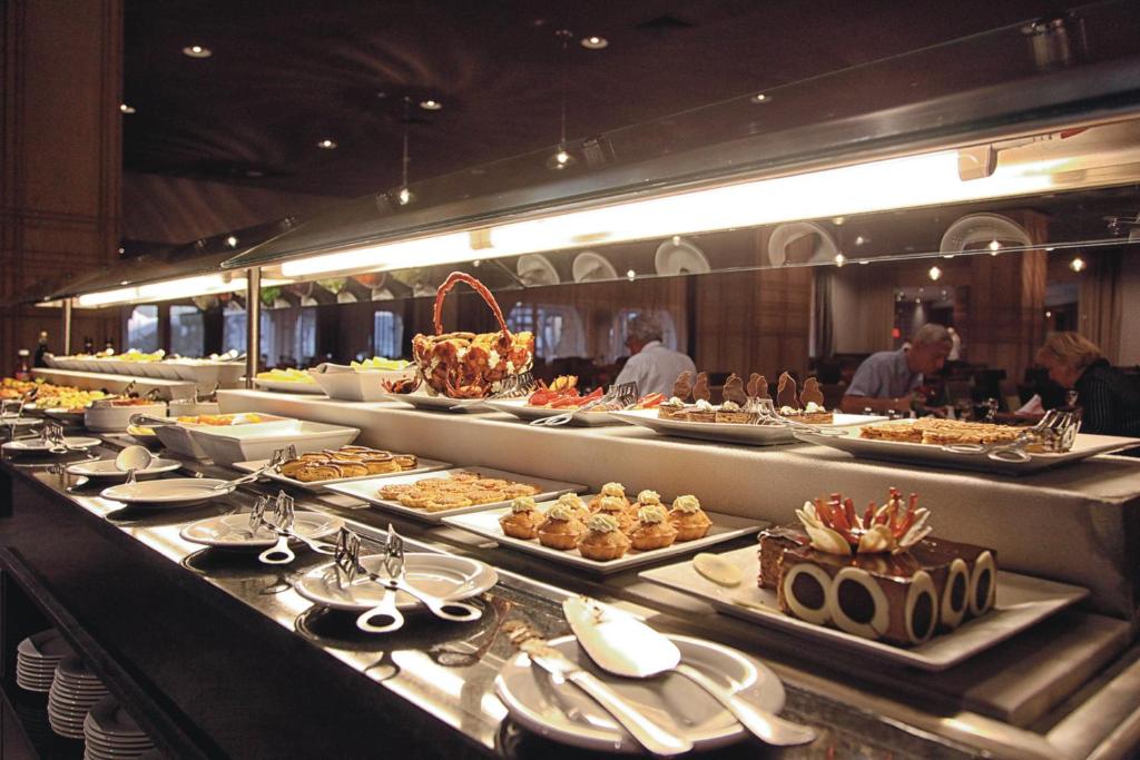 Bufete ar plašu dažādu ēdienu izvēli, tostarp saldējumus un uzkodas, kas sakārtotas uz galda ar sudraba traukiem un šķīvjiem. Gaisotne ir eleganta un viesmīlīga.