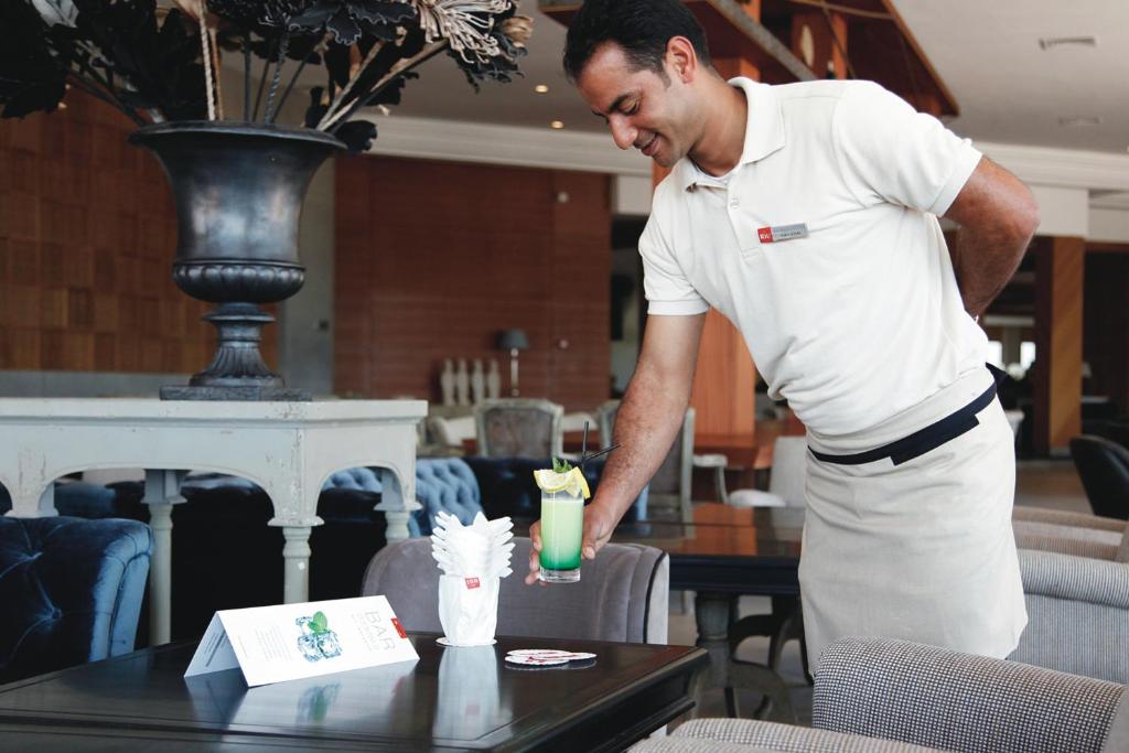Viesmīlis pasniedz kokteili restorānā, kam blakus ir ēdienkarte un salvetes. Interjers ir elegants ar mīkstiem krēsliem un dekoratīviem elementiem.