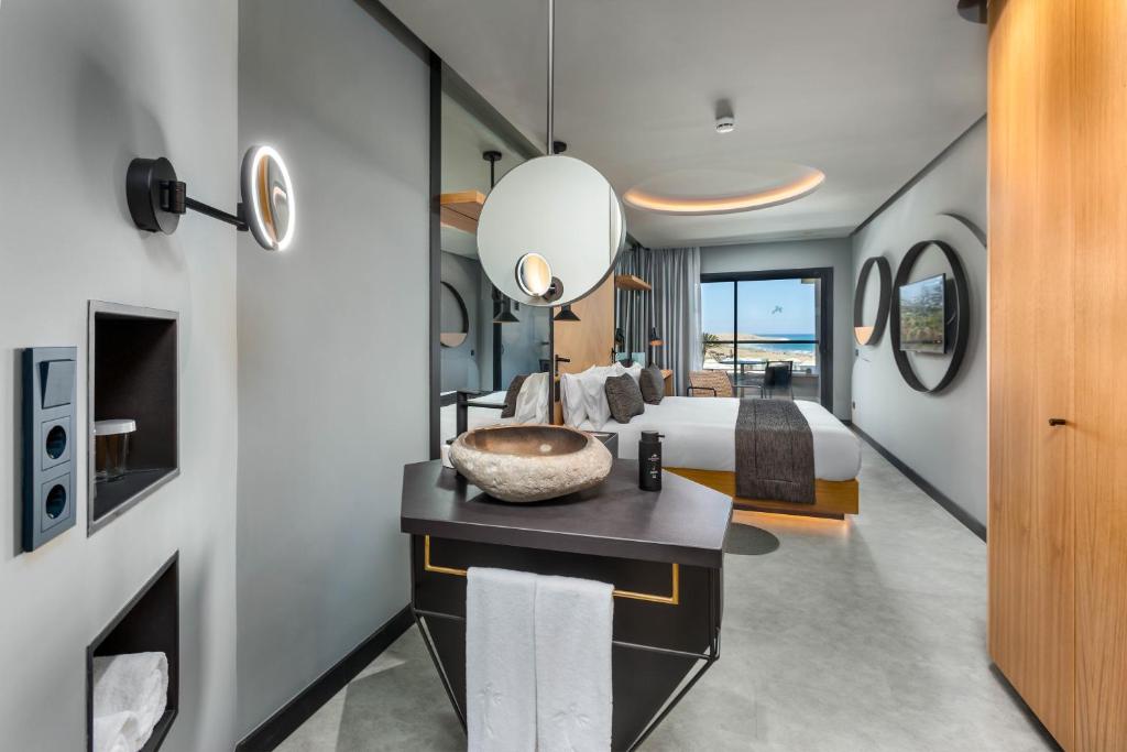 Moderns viesnīcas numurs ar elegantu interjeru, kas ietver gultu, dekoratīvos spoguļus un skatu uz pludmali. Numurā redzams arī stilīgs izlietnes galds un mūsdienīgas apgaismojuma ierīces.