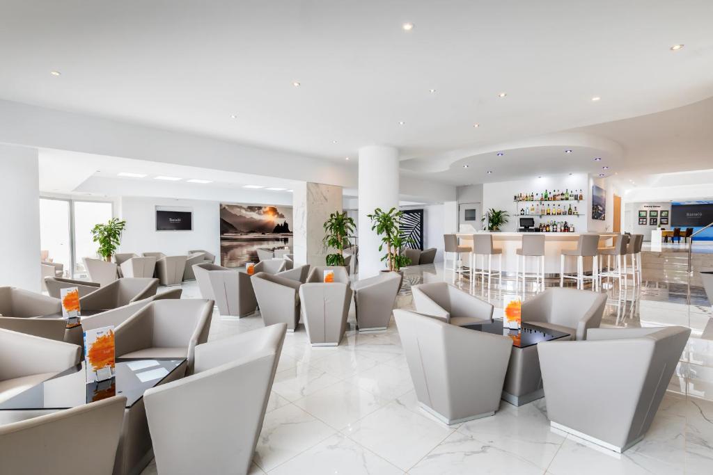 Moderns bārs ar elegantiem krēsliem un galdiem, kas veidots gaišā interjerā. Telpā ir daudz dabīgas gaismas un zaļo augu, radot relaksācijas atmosfēru.