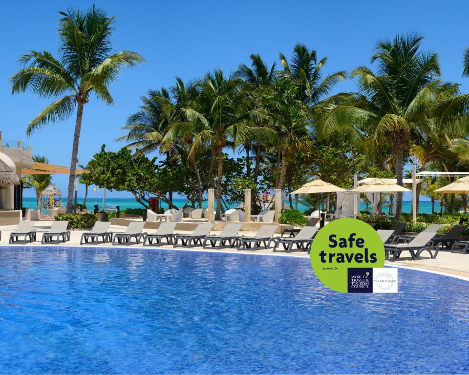 Tropu kūrorts ar baseinu un palmām, kas atrodas tuvu pludmalei, ar zīmi "Safe travels".