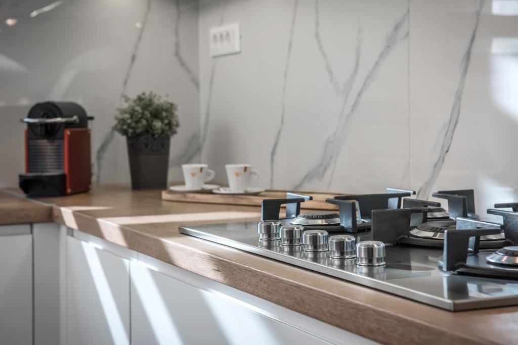 Mūsdienīga virtuve ar gāzes plīti, koka virsmas un elegantu kafijas automātu, kas papildina interjeru. Uz galda redzami balti krūzītes ar zeltītiem akcentiem.