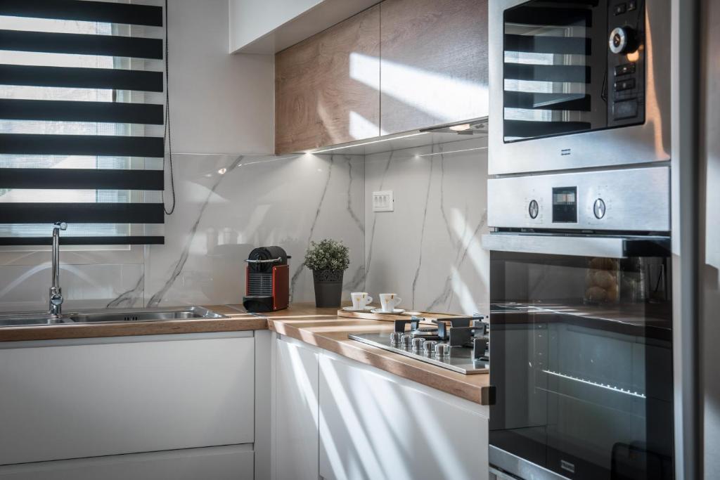 Mūsdienīga virtuve ar gaismas caurlaidīgiem logiem, melnām un baltām žalūzijām, izsmalcinātu sienu apdari un modernu virtuves tehniku. Uz galda atrodas kafijas automāts un neliels puķu pods.