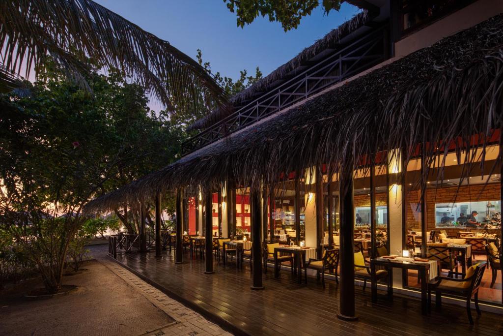 Tropu stila restorāns ar salmu jumtu, apgaismots pie vakara saulrieta. Terase ar galdiem un krēsliem, kas ierāmēta ar zaļām augiem.