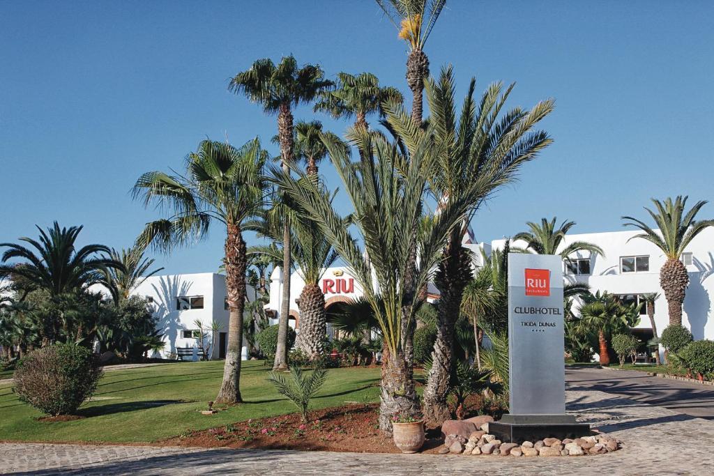 Zaļš dārzs ar palmām un zālienu pie Riu Clubhotel Tikida Dunas ieejas, kur redzams viesnīcas nosaukums.
