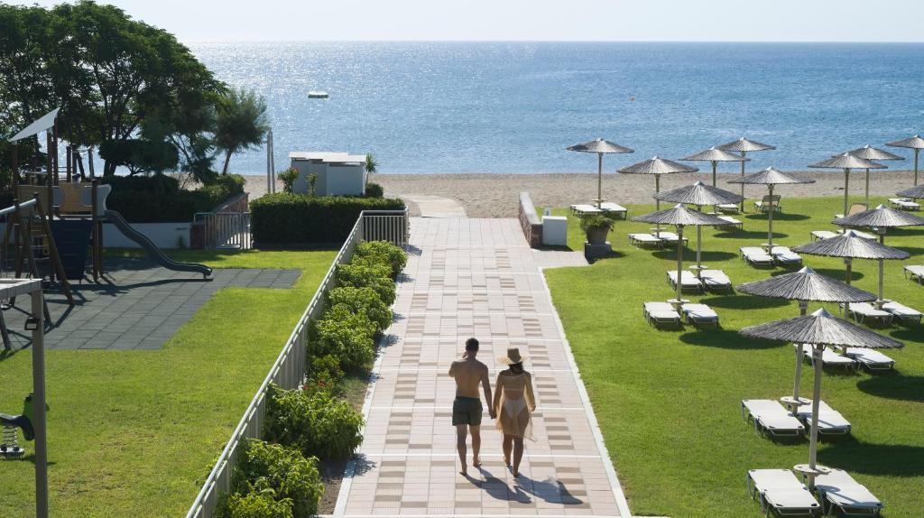 Pāris pastaigā pa bruģētu celiņu, kas ved uz pludmali ar saulessargiem un saulē apsildītām gultām. Jūra mirdz saulē, radot mierīgu un relaksējošu atmosfēru.