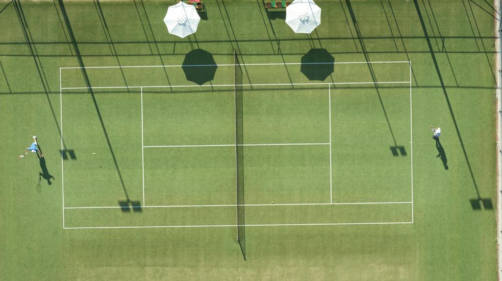 Divi tenisisti spēlē uz zaļā laukuma, apgādāta ar saules parasoliem. Skats no augšas parāda laukuma līnijas un ēnu veidojumus.