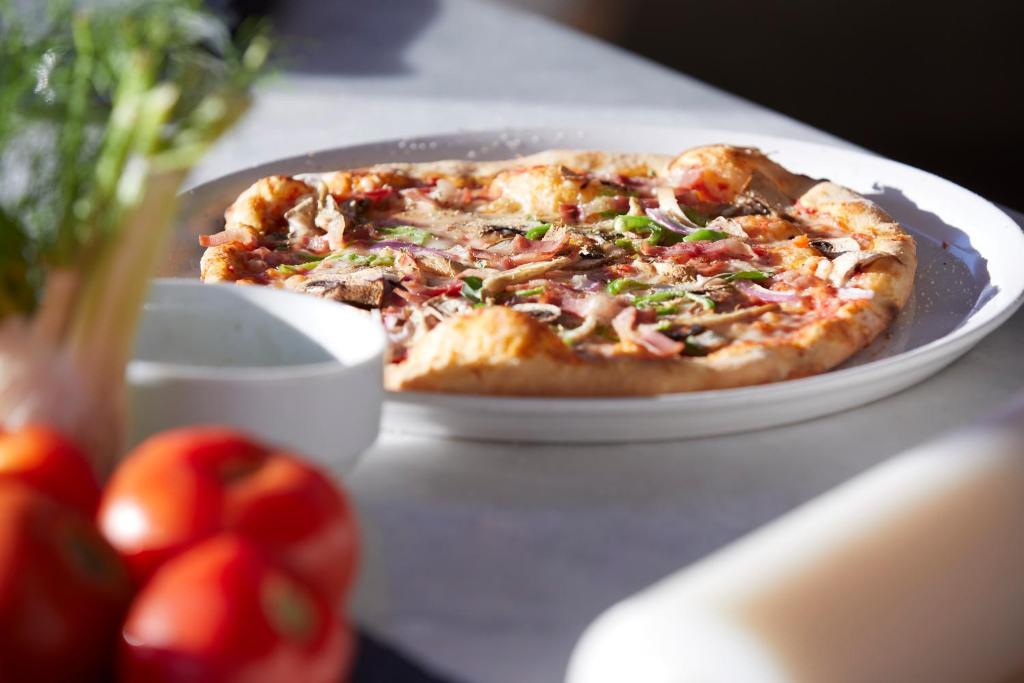 Svaiga pica ar gaļu, dārzeņiem un siera pārklājumu uz balta šķīvja, blakus tomātiem un mērci.