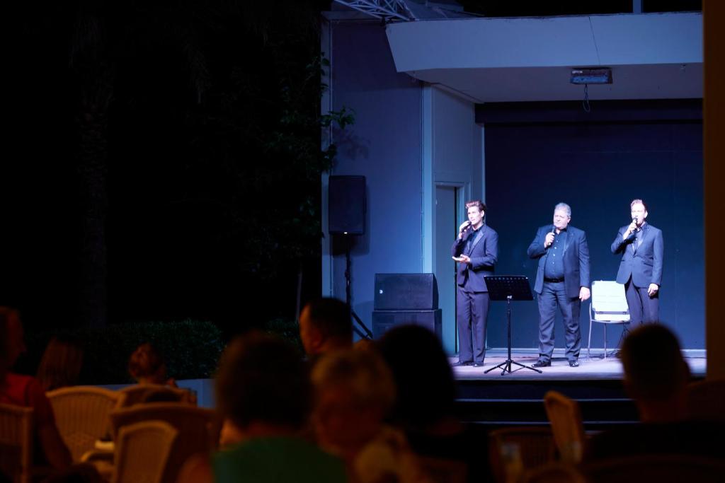 Trīs dziedātāji uz skatuves, sniedzot priekšnesumu tumšā vidē, kamēr apmeklētāji bauda izrādi.