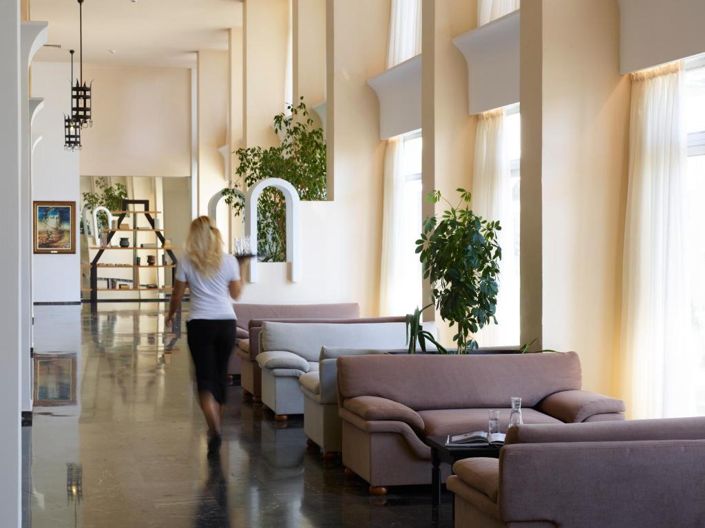 Mūsdienīgs viesnīcas interjers ar gaišiem toņiem, plašām telpām un augstiem logiem, kurā redzama sieviete, kas pārvietojas pa telpu. Dabas elementi un eleganta mēbele rada relaksējošu atmosfēru.