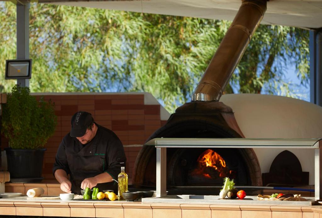 Šefpavārs gatavo ēdienus pie atvērtas virtuves ar koka krāsni, kurā deg uguns. Uz galda ir svaigi dārzeņi un olīveļļa, radot viesmīlīgu un radošu atmosfēru.