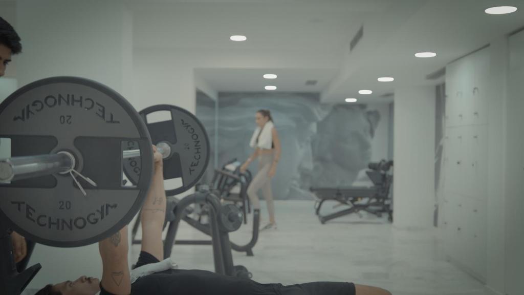 Sporta zālē notiek treniņš, kur vīrietis paceļ svarus, kamēr blakus stāv sieviete. Telpa ir moderna un gaiša, ar sporta aprīkojumu un atslēgu skapīšiem.