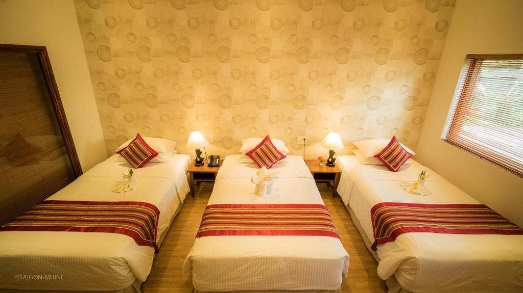 Mūsdienīga viesnīcas istaba ar trim atsevišķām gultām, dekorētām ar sarkanām spilvendrānām. Istabā ir naktslampas un koka galdiņš, kas piepildīts ar mājīgām detaļām.