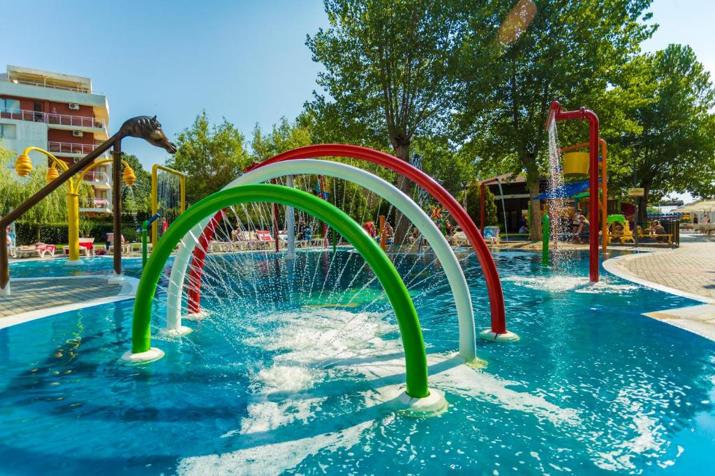 Krāsains ūdens atrakciju parks ar strūklakām un rotaļu elementiem, ideāls vieta bērniem izklaidēties karstā vasaras dienā. Apkārt redzami koki un atpūtas zonas.