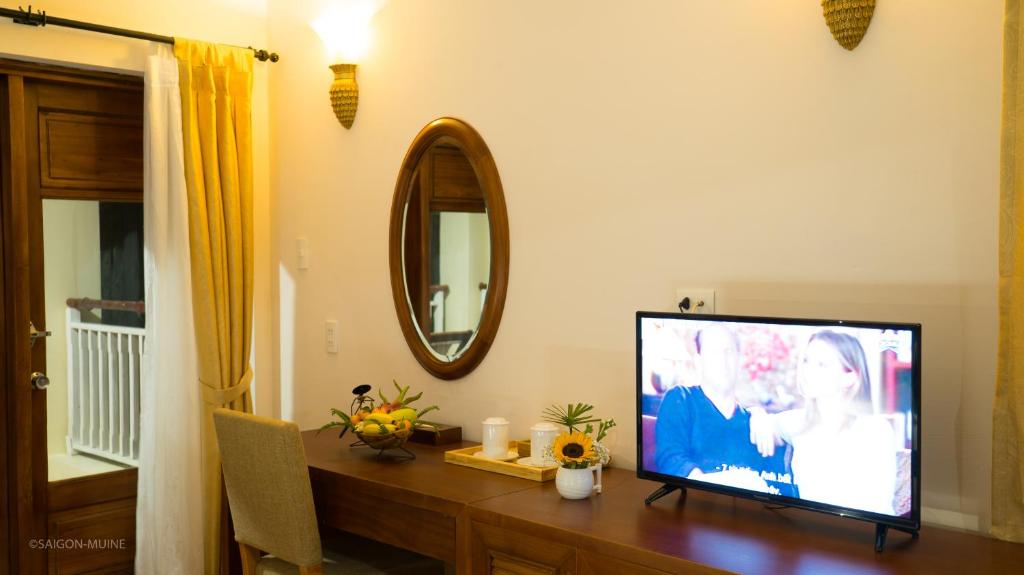 Mūsdienīgs viesnīcas numurs ar televizoru, rakstāmgaldu un spoguli, kas atspoguļo elegantu interjeru ar dzeltenām aizkariem un augļiem uz galda.