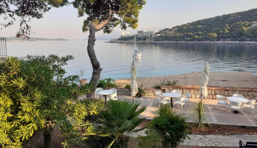 Rāmja skats uz mierīgu piekrastes teritoriju ar krastā esošām baltiem galdiem un krēsliem. Fons ietver jūru ar laivu un zaļajiem pakalniem.