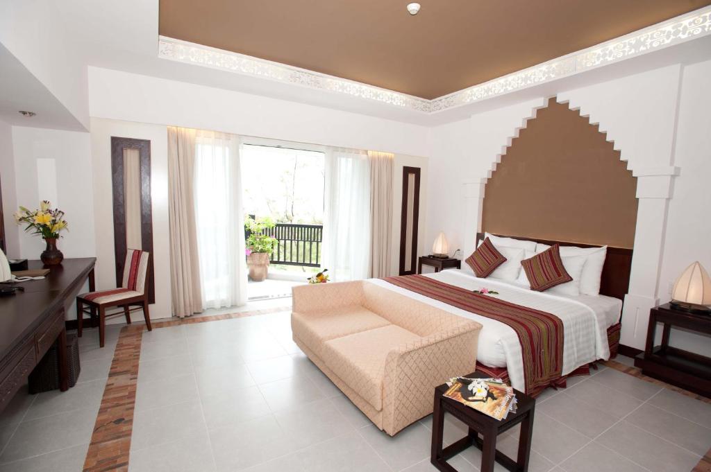 Mūsdienīga viesnīcas numura iekārtojums ar ērtu gultu, dīvānu un lielām logu durvīm, kas ved uz terasi. Interjers ir elegants ar siltām krāsām un dekoratīviem elementiem.