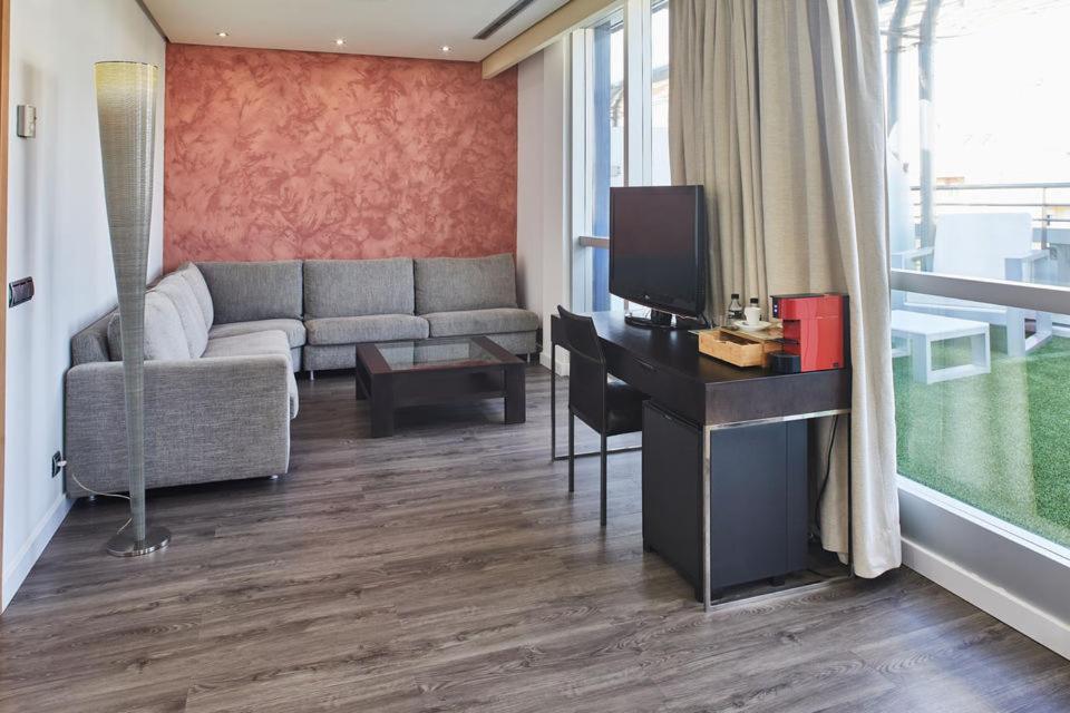 Mūsdienīga viesnīcas numura interjers ar pelēku dīvānu, stikla galdu un televizoru, ko papildina siltas krāsas sienas un gaišas aizkari. 