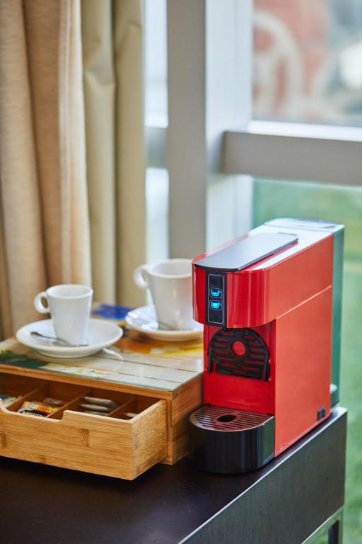 Sarkana kafijas automāts blakus divām baltām kafijas tasītēm un bambusa trauku kastei ar kafijas piedevām.