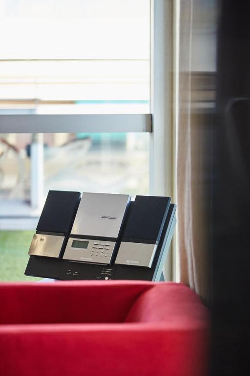 Mūzikas sistēma ar melniem skaļruņiem un metāla apdari, novietota pie loga, blakus sarkanai dīvāna malai.