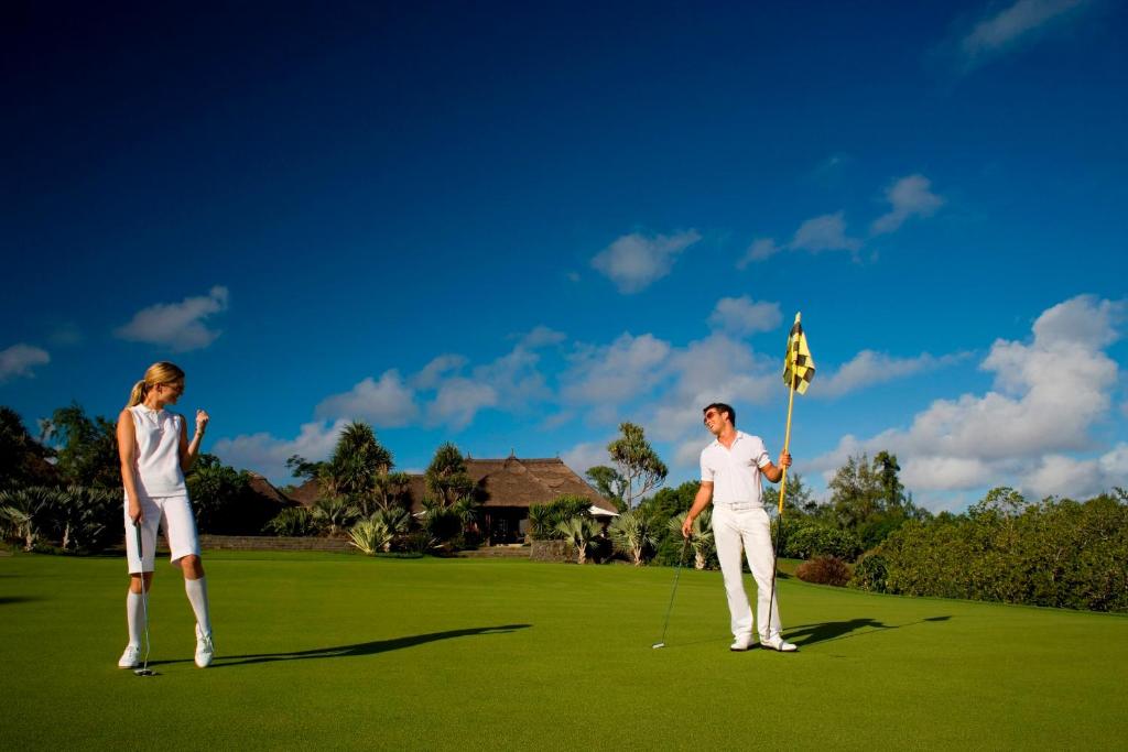 Divi golfa spēlētāji, ģērbušies baltos tērpos, spēlē uz zaļa golfa laukuma, ar skaidru zilu debesu fona. Vīrietis tur karogu, kamēr sieviete vēro bumbu.