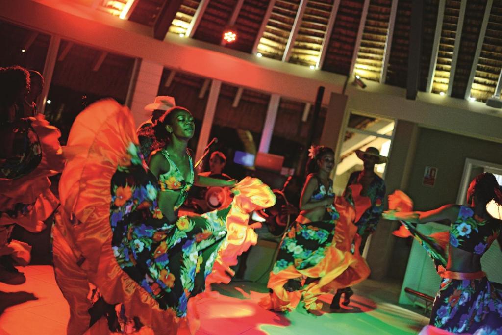 Dejotāju grupa, kas tērpta krāsainās, ziedu motīvu tērpos, veic enerģisku un dinamisku deju zem sarkano un zaļo gaismu apgaismojuma.