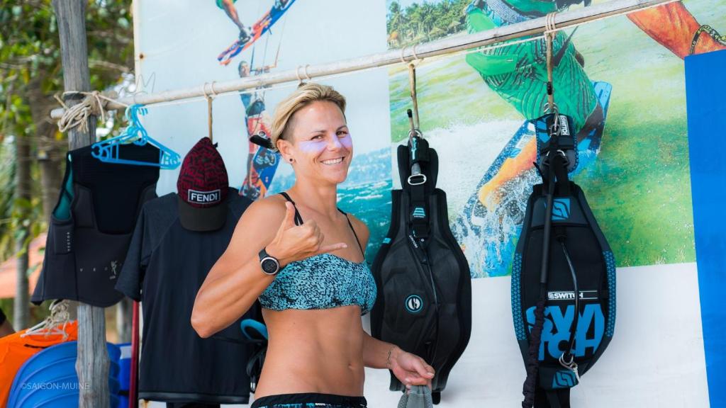 Sieviete pludmalē ar sporta apģērbu, parādot "OK" zīmi, fona attēlā redzama ūdens sports aktivitāte. Apģērbi un aprīkojums karājas pie stenda.