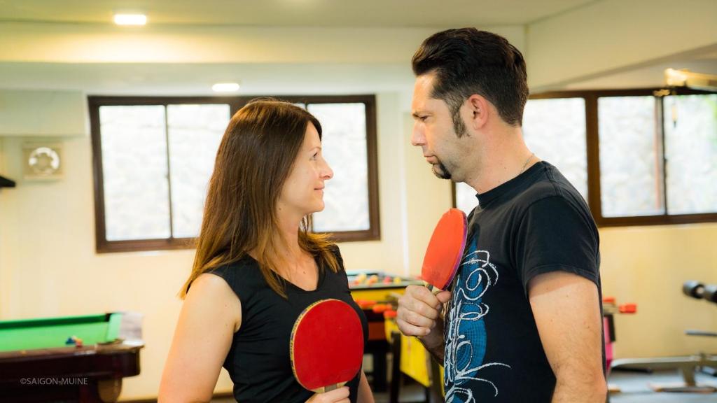 Divi cilvēki skatās viens otram acīs, turēt pingpongu raketes rokās. Fons ir sporta zāle ar dažādām spēļu galdiem un logiem.