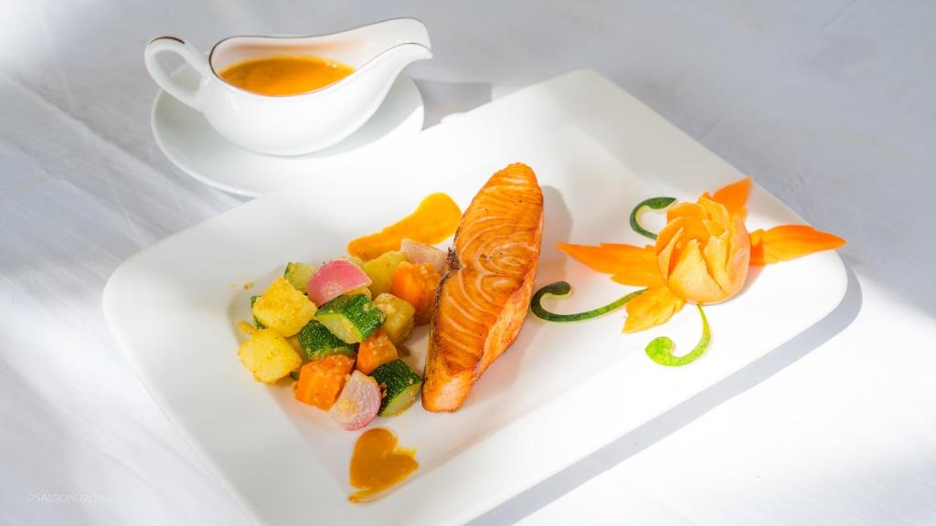 Garšīgs cepts lasis ar dārzeņu salātiem un dekoratīvu burkānu ziedu uz balta šķīvja, blakus ir mērces trauks.