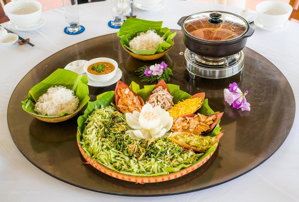 Skaista galda kompozīcija ar dažādiem ēdieniem, tostarp rīsu nūdelēm, garnelēm un dārzeņiem, dekorēta ar ziediem un pasniegta uz apaļa galda.