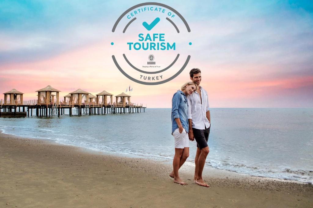 Pāris pastaigā pa pludmali pie saulrieta, fonā redzami koka paviljoni un "Safe Tourism" sertifikāts.