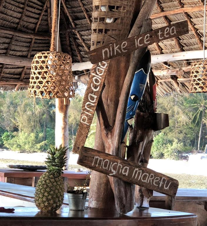 Kokteiļu bāra stends ar zīmēm, kas norāda uz "Pina Colada", "Nike Ice Tea" un "Maicha Marefu", pie galda ar ananāsu un dabisku ainavu fona.