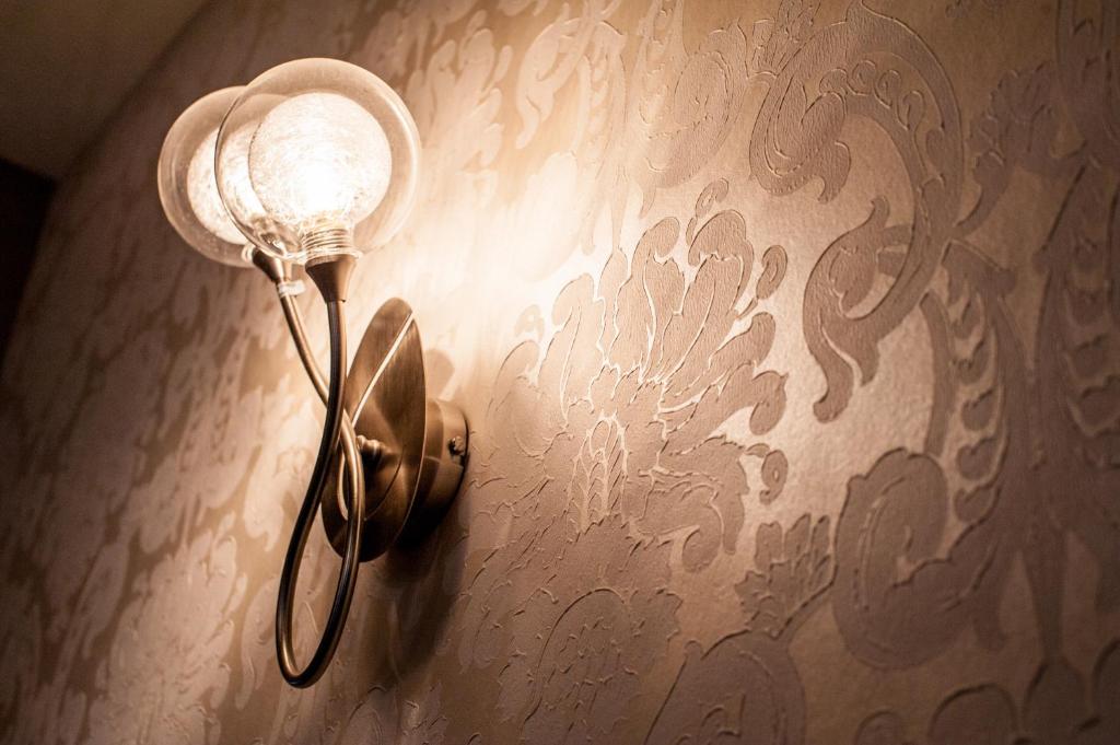 Sienas lampa ar divām stikla spuldzēm, kas izgaismo elegantu reljefotu tapeti ar florālu rakstu.