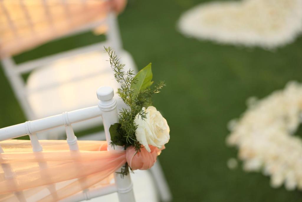 Kāzu dekorācija ar baltu ziedu un zaļām lapām, kas piestiprināta pie baltas krēsla atzveltnes, ar maigu persiku krāsas lentu. Fons ir zaļš zāliens ar ziedu izkārtojumu.