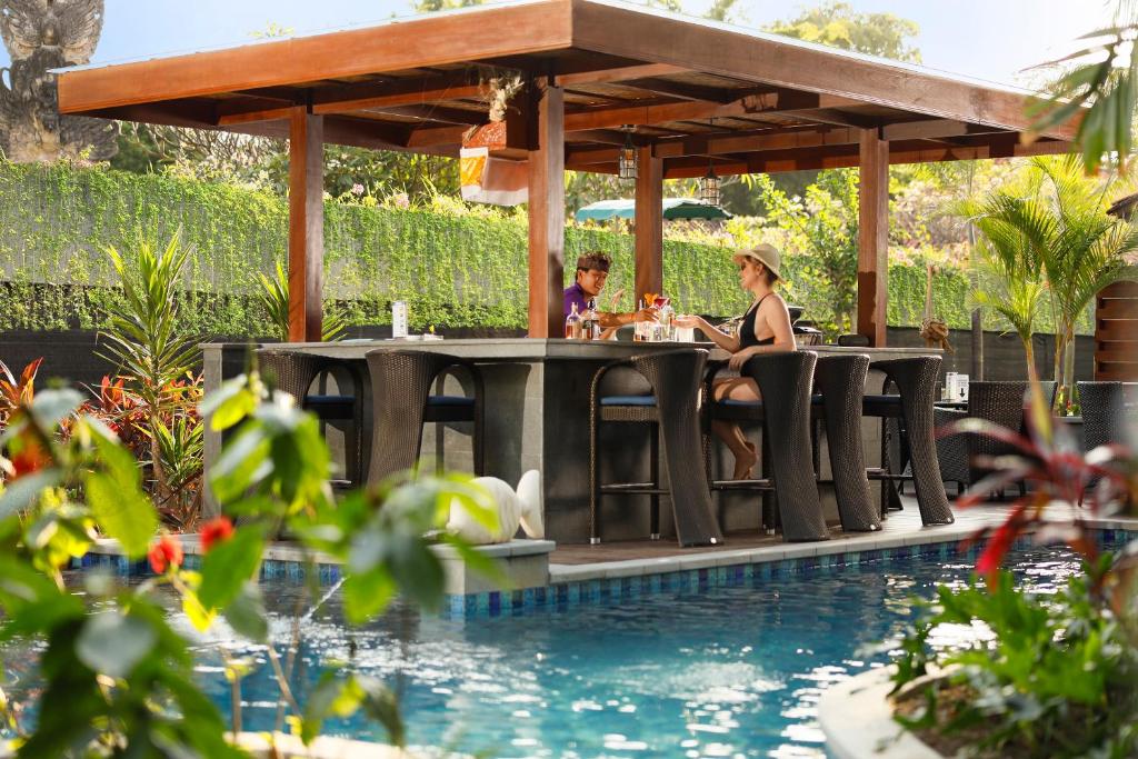 Divi cilvēki sēž pie bāra galda zem nojumes blakus baseinam, baudot dzērienus un vieglas uzkodas. Apkārt ir sulīgs zaļums un krāsaini ziedi, radot relaksējošu atmosfēru.