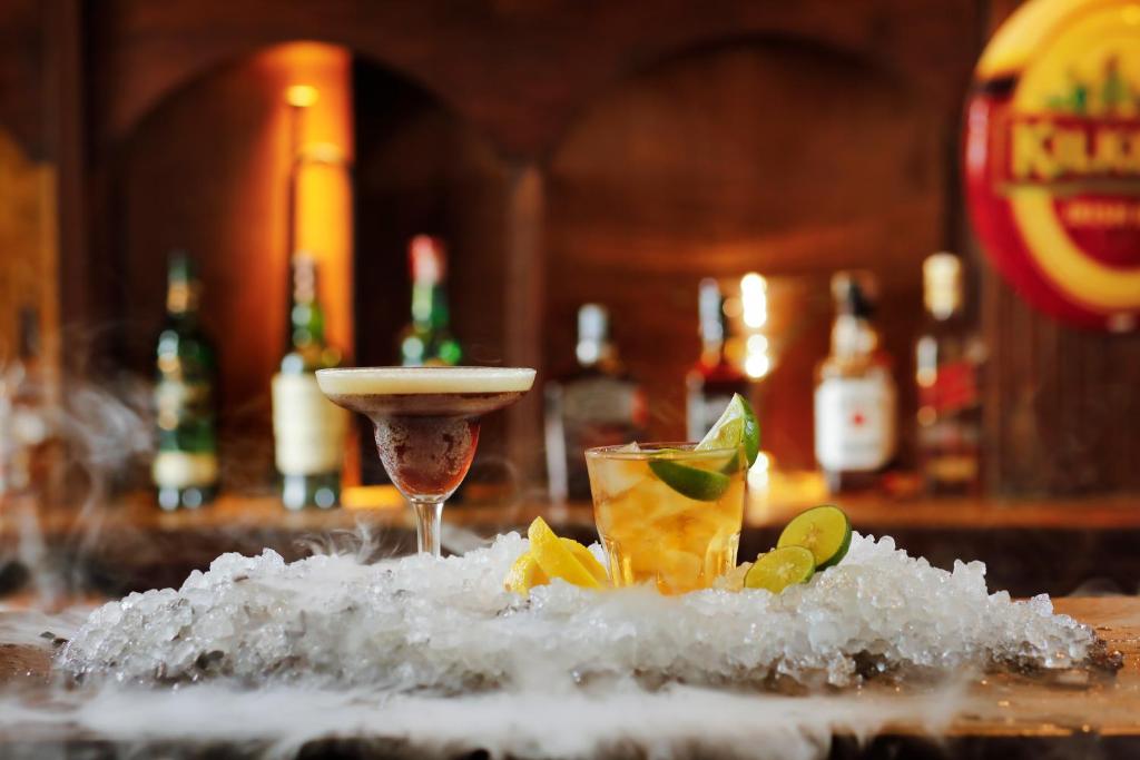 Divi kokteiļi uz ledus, viens ar putu virsotni un otrs ar laima šķēlēm, fona apgaismojums un bāra dzērieni.