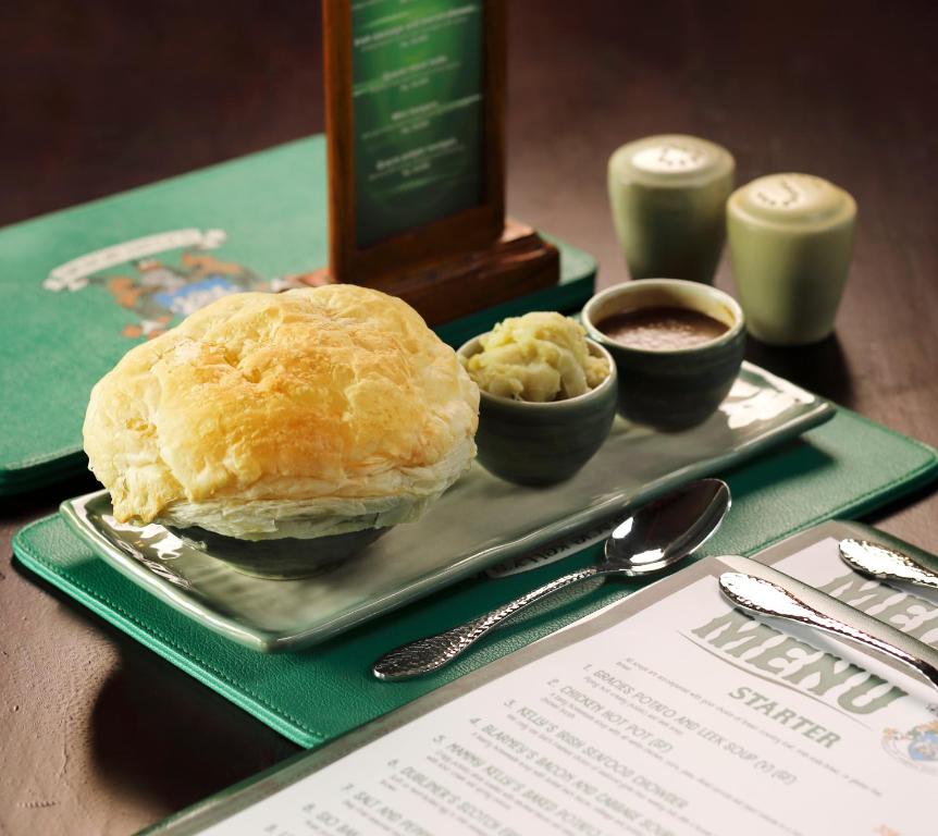 Svaiga cepta pīrāga porcija ar mīkstu pildījumu un divām mērču bļodiņām, pasniegta uz elegantas plāksnes kopā ar sānu ēdienu. Fonā redzams ēdienkarte un galda piederumi.