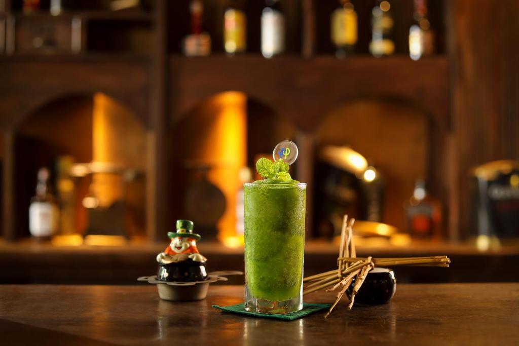 Zaļš kokteilis ar piparmētru un dekoratīvu elementu uz ledus, novietots uz koka galda blakus maziem priekšmetiem. Fons ar izsmalcinātām dzērienu pudelēm rada siltu un viesmīlīgu atmosfēru.