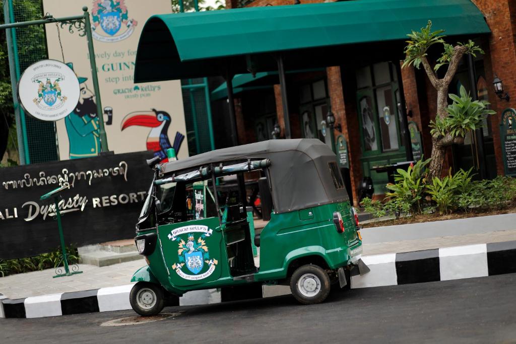 Zaļš tuk-tuks stāv pie Bali Dynasty Resort ieejas, fonā redzama viesnīcas zīme un augsne ar sulīgām augiem.