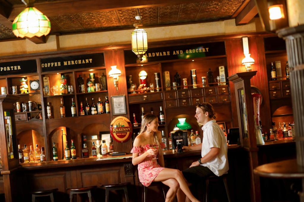 Bāra ainas, kur divi cilvēki sēž pie letes, baudot dzērienus un sarunājoties. Aiz viņiem redzama bāra plauktu rinda ar dažādām alkoholiskajām dzērienu pudelēm.