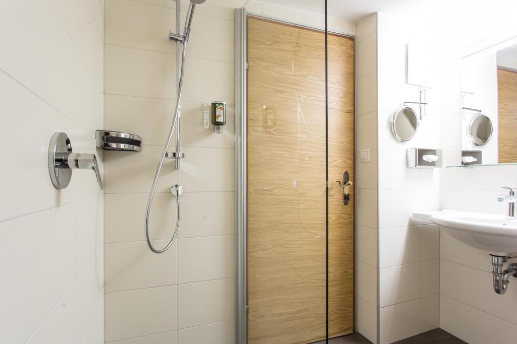 Mūsdienīgs vannas istabas interjers ar stikla dušu, hroma dušas sistēmu un koka durvīm. Kvalitatīvs apgaismojums un tīras līnijas rada elegantu atmosfēru.